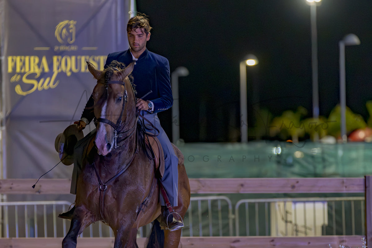 A man in a dark navy blue velvet equestrian jacket rides a brown horse during an equestrian event at night under stadium lights, with a banner reading 'FEIRA EQUESTRE Sul' in the background.