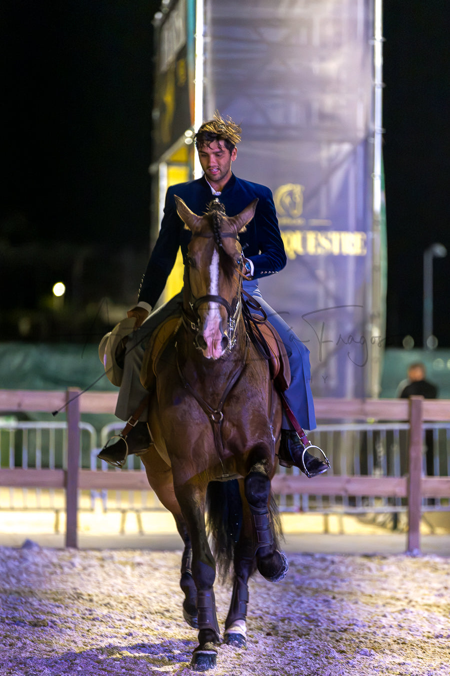 A man in formal equestrian Portuguese Traditional attire rides a brown horse in an outdoor night arena, with a large illuminated banner and fencing visible in the background.