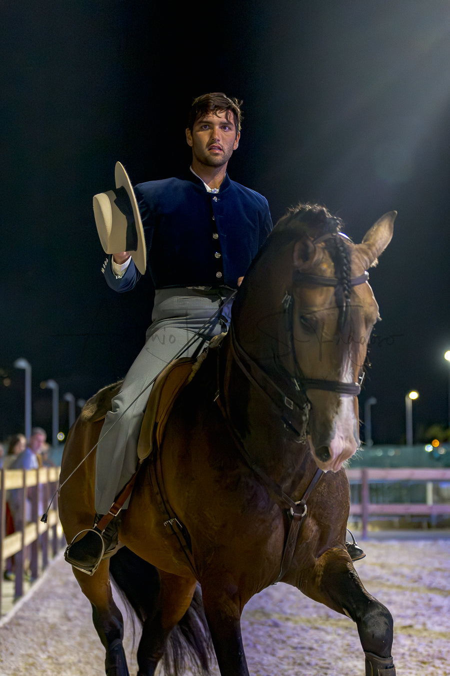 A man in a dark blue historical military-style jacket and light gray trousers rides a brown horse in a night arena, holding a light-colored Portuguese Traditional hat in one hand.