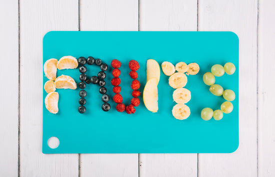 Fruits arranged on a teal cutting board to spell out 'FRUITS' on a white wooden background