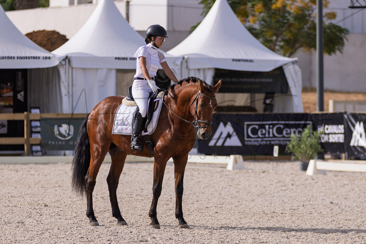 A rider in white equestrian attire sits on a brown horse in a sandy riding arena, with white event tents and branded barriers in the background.