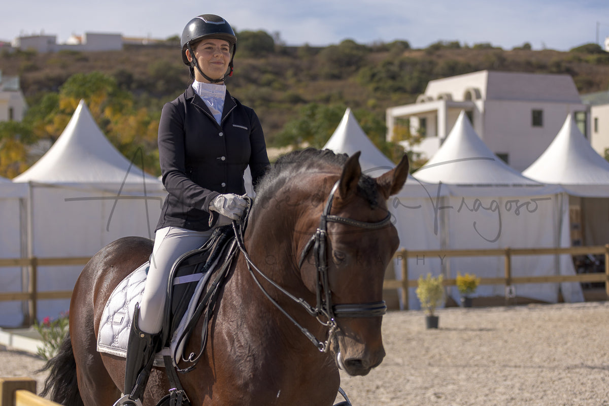 A woman in equestrian attire sits on a dark brown horse in an outdoor riding arena, with white event tents and hills in the background.