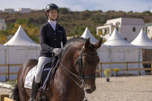 A woman in equestrian attire sits on a dark brown horse in an outdoor riding arena, with white event tents and hills in the background.